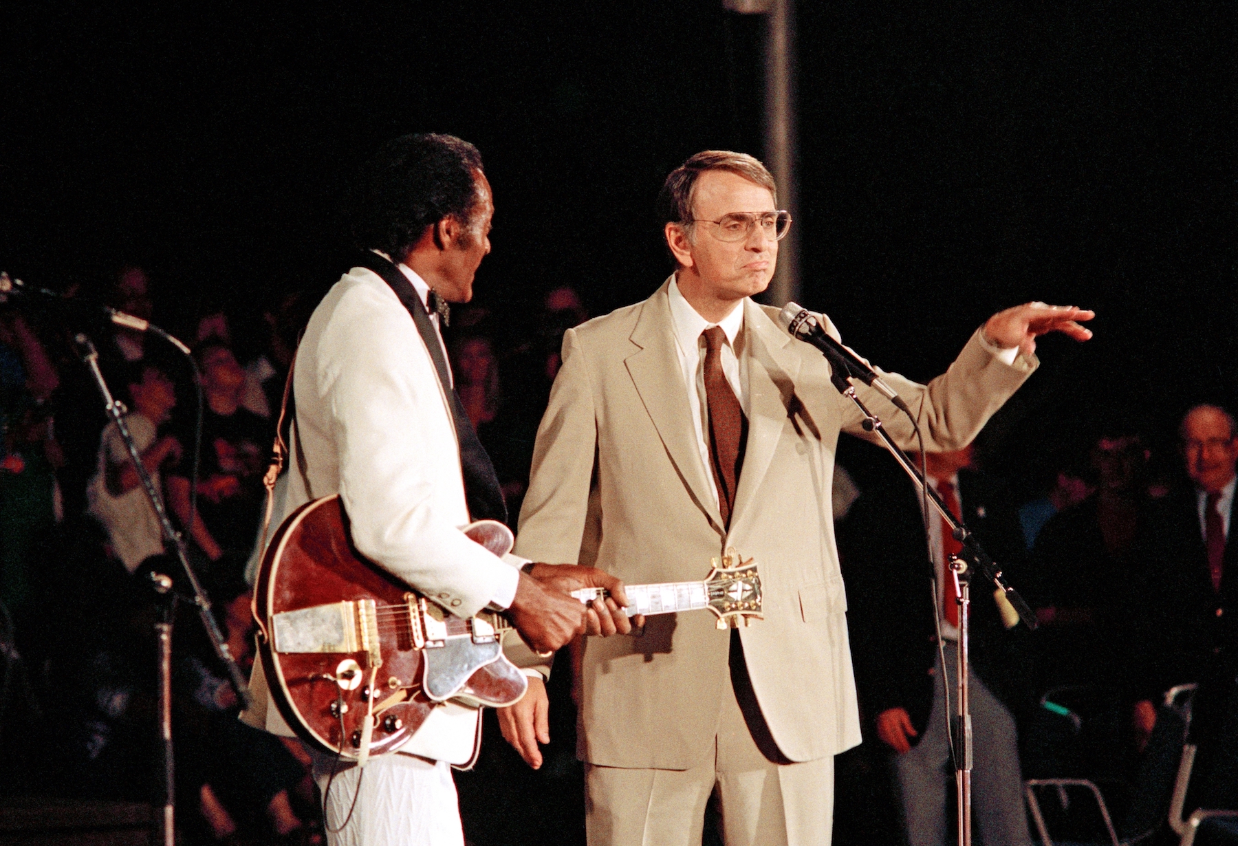 man holding guitar and another man gesturing standing at microphone