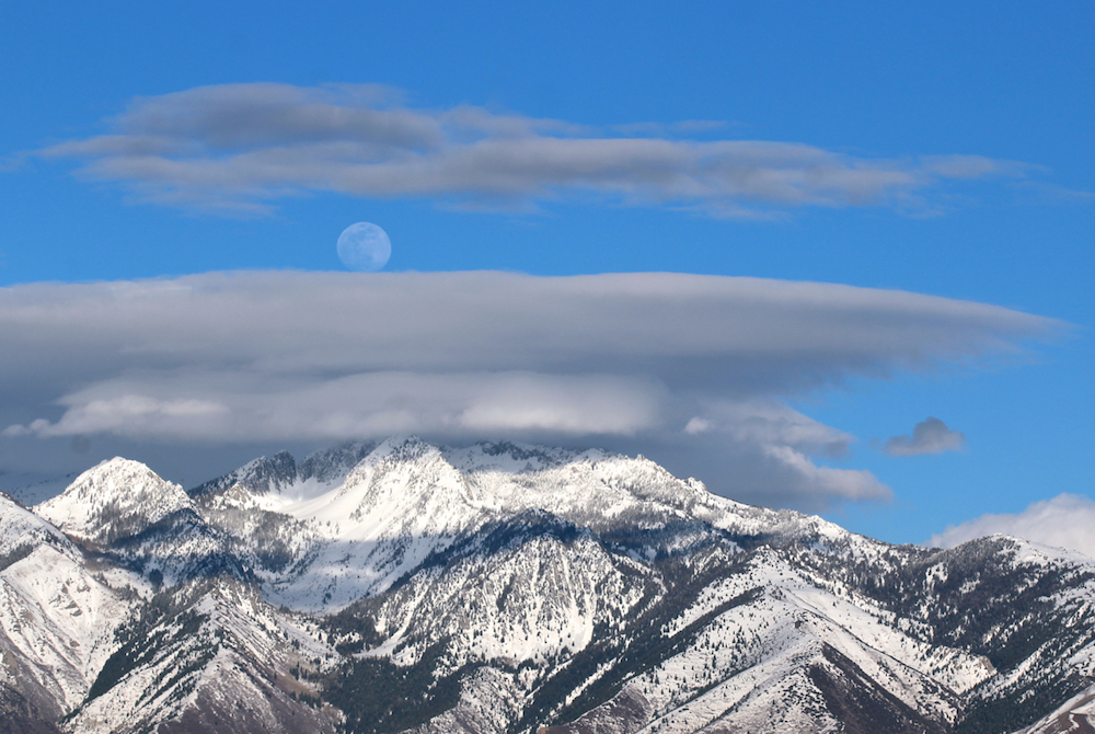 daylight moon over lenticular clouds and snowy mountaintops